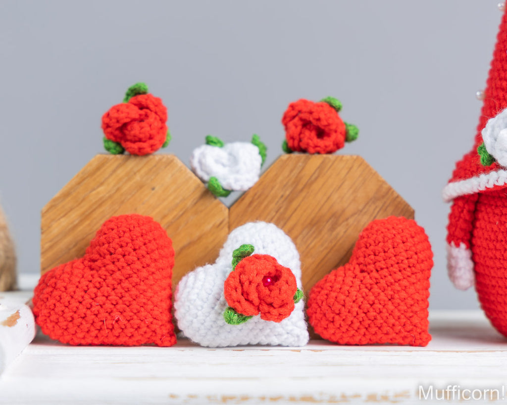 Handmade crocheted decorative items including red and white hearts, small wooden house-shaped stands, and red roses with green leaves, arranged on a white surface against a light gray background.