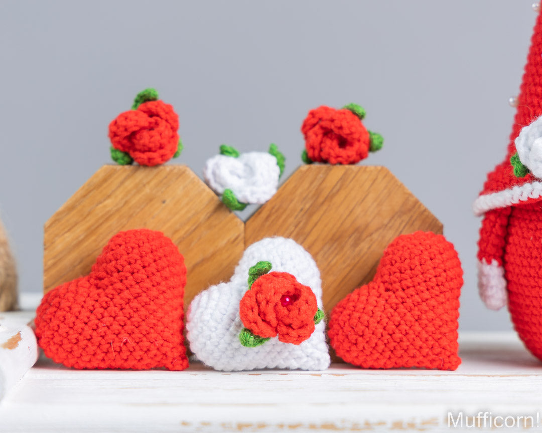 Handmade crocheted decorative items including red and white hearts, small wooden house-shaped stands, and red roses with green leaves, arranged on a white surface against a light gray background.