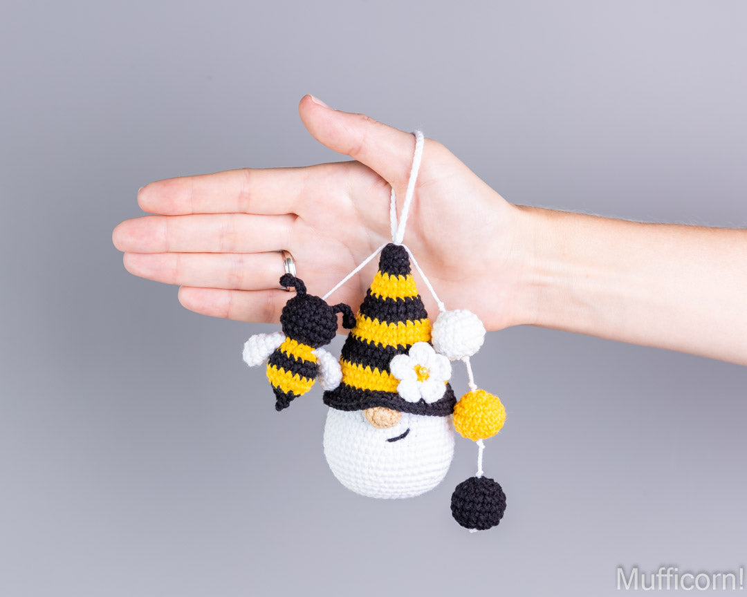 A hand holding a crocheted bumblebee and gnome keychain against a solid gray background.