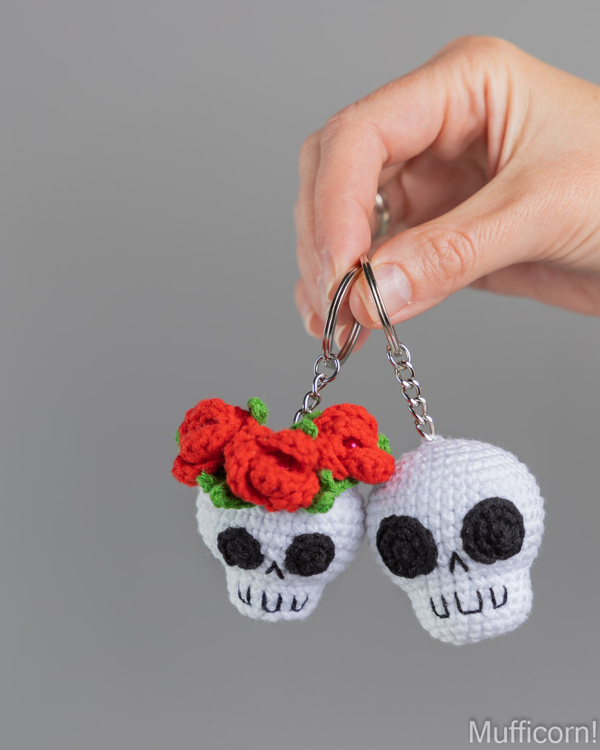 A hand holds two crocheted skull keychains against a plain gray background. One skull has red flowers with green leaves, and the other is plain white with black embroidered facial details.