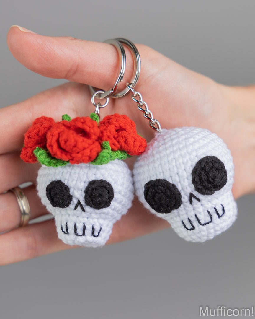 A hand holds two crocheted skull keychains against a plain gray background. One skull is white with black embroidered facial features and red crocheted roses on top with green leaves; the other is plain white.