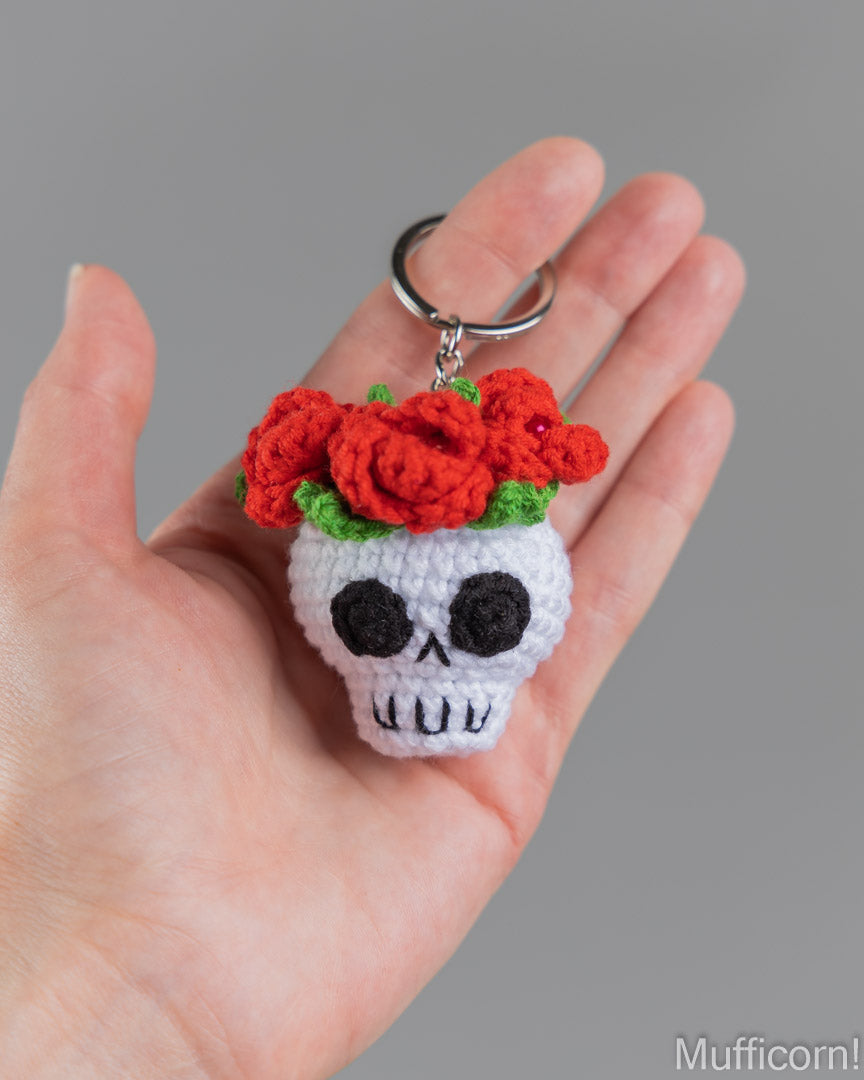 A crocheted sugar skull keychain with red roses, held in a person's hand against a solid gray background.