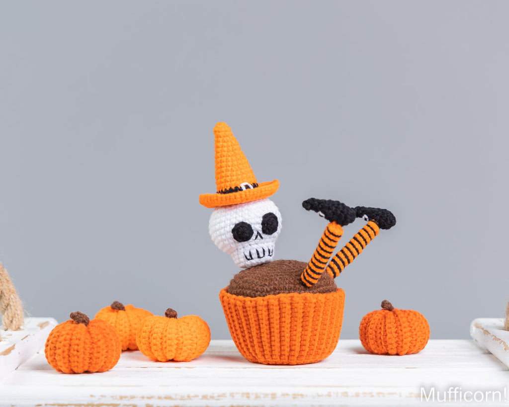 A crocheted Halloween-themed cupcake decoration featuring a white skull wearing an orange witch hat, sitting in an orange cupcake liner with brown frosting and striped orange-and-black legs. Small crocheted pumpkins are arranged around it on a white surface against a light gray background.