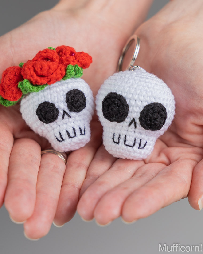 Two crocheted skull keychains held in a person's hands. One skull is white with black embroidered facial features and a crown of red roses with green leaves; the other is plain white with black facial features.
