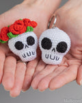 Two crocheted skull keychains held in a person's hands. One skull is white with black embroidered facial features and a crown of red roses with green leaves; the other is plain white with black facial features.