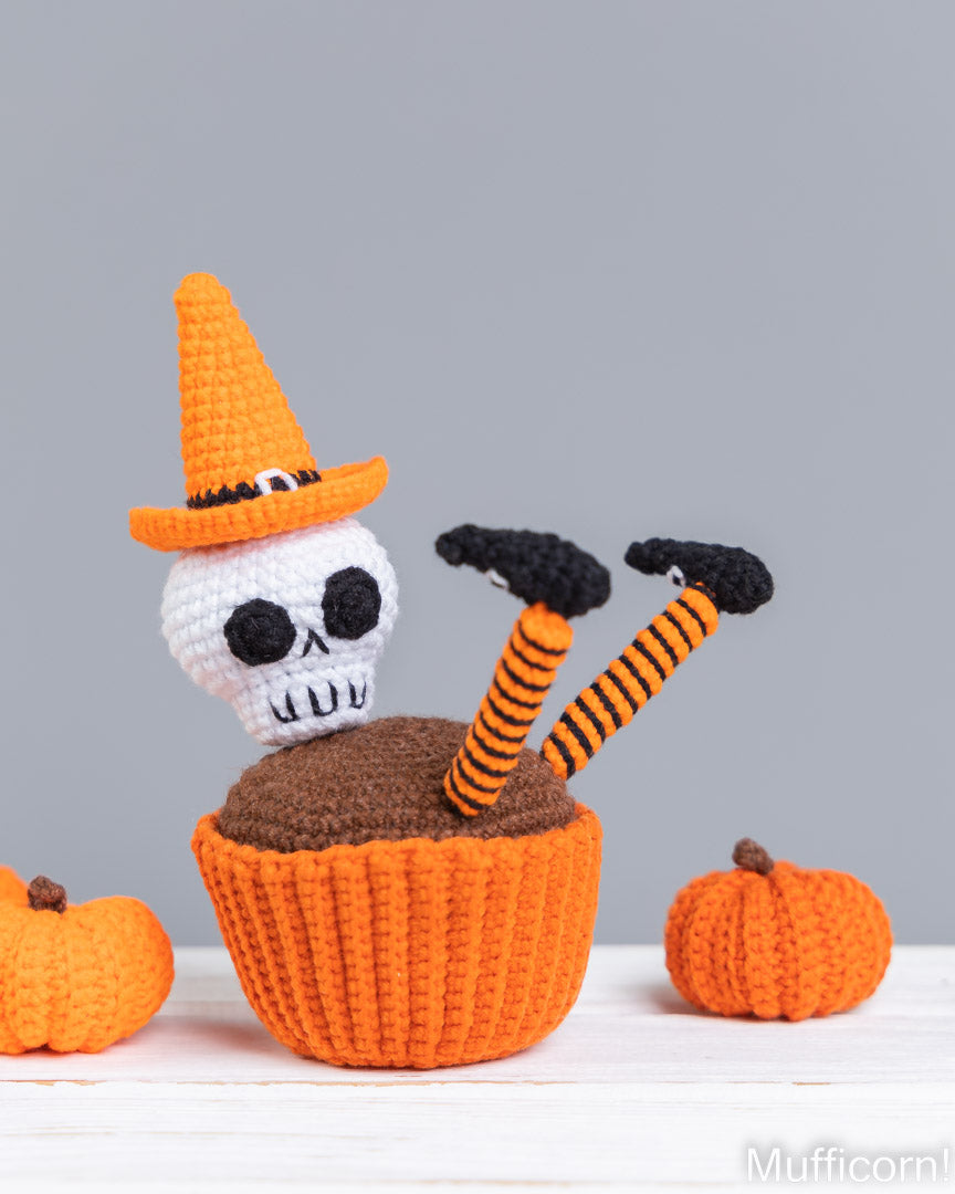 A crocheted Halloween-themed cupcake decoration featuring a white skull wearing an orange witch hat, with orange-and-black striped legs sticking out of a brown cupcake liner, set on a white surface with small orange pumpkins against a gray background.