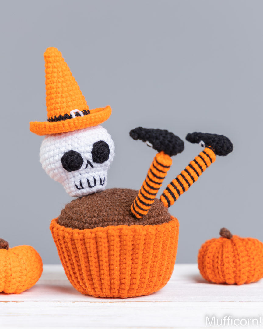 A crocheted Halloween cupcake decoration featuring a white skull wearing an orange witch hat, rising from an orange cupcake liner with orange-and-black striped legs, shown with small pumpkin props on a gray background.