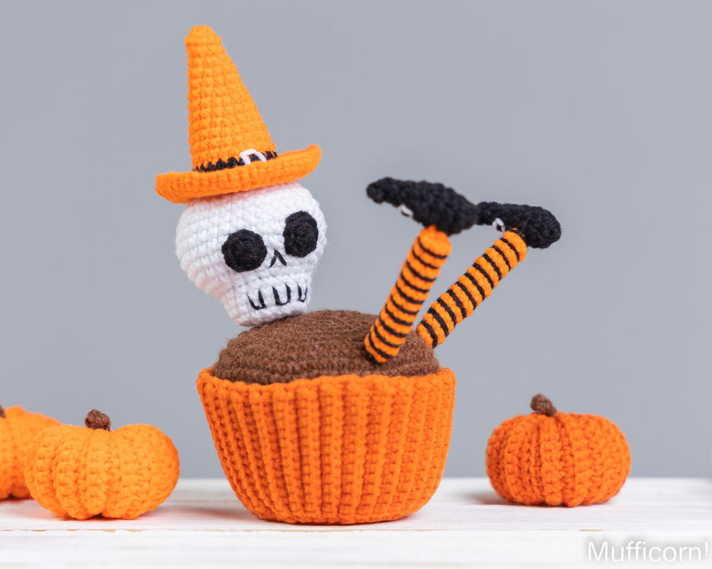 A crocheted Halloween-themed amigurumi decoration featuring a white skull wearing an orange witch hat, emerging from an orange cupcake with brown frosting and striped orange-and-black legs. Small orange pumpkins sit on a white surface against a light gray background.