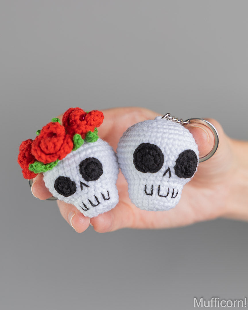 A hand holding two small crocheted skull keychains against a plain gray background. One skull is white with black embroidered details and a red flower arrangement on top, and the other is plain white with black details.