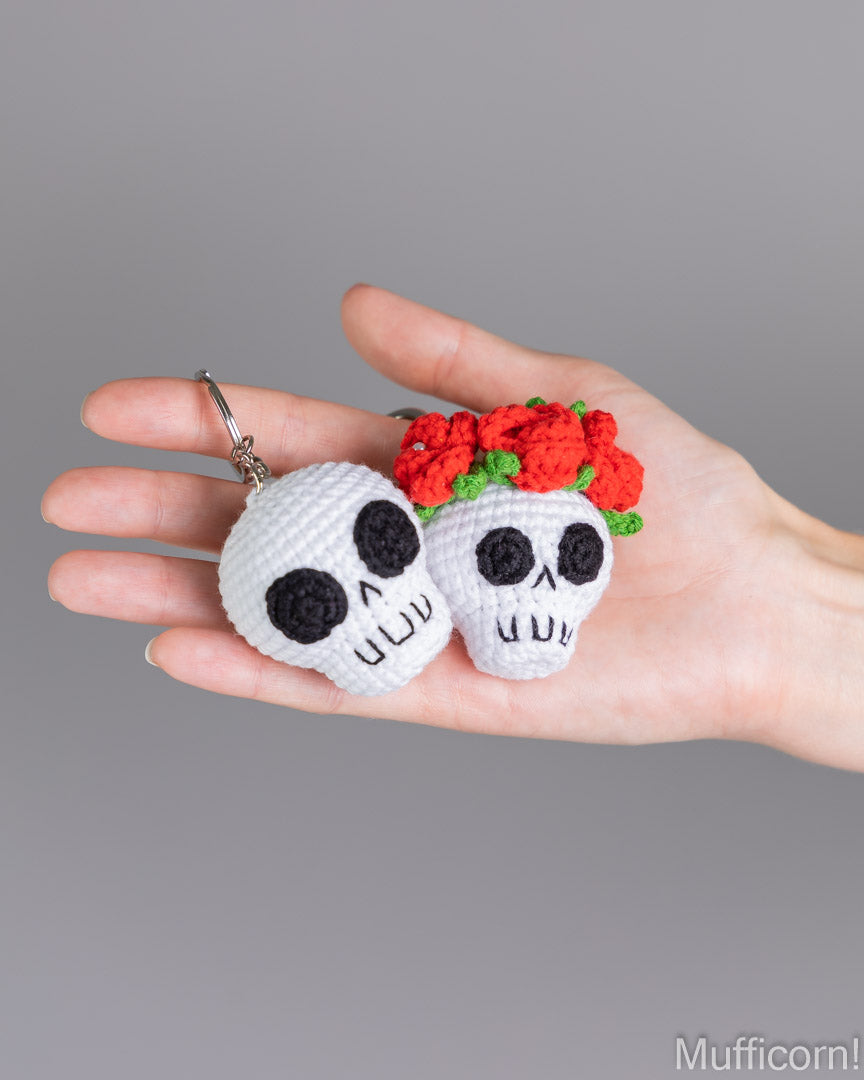 Two white crocheted sugar skull keychains are held in a person's hand against a neutral gray background. One skull has black yarn details, and the other has a red and green crocheted flower crown.