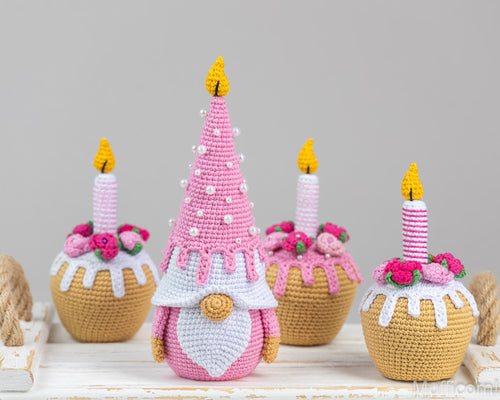 Three crocheted gnome cupcake decorations arranged on a white wooden surface against a light gray background. The central gnome is pink with a white chest, yellow flame candle, and pink-and-white striped candle.