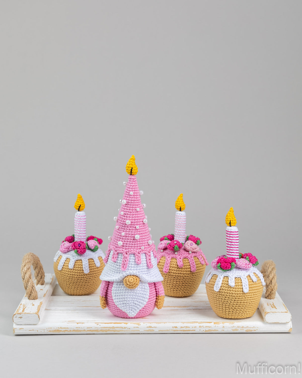 A crocheted holiday display featuring a central pink gnome with a pointed hat and three cupcake-like figures with candles, arranged on a white tray against a light gray background.