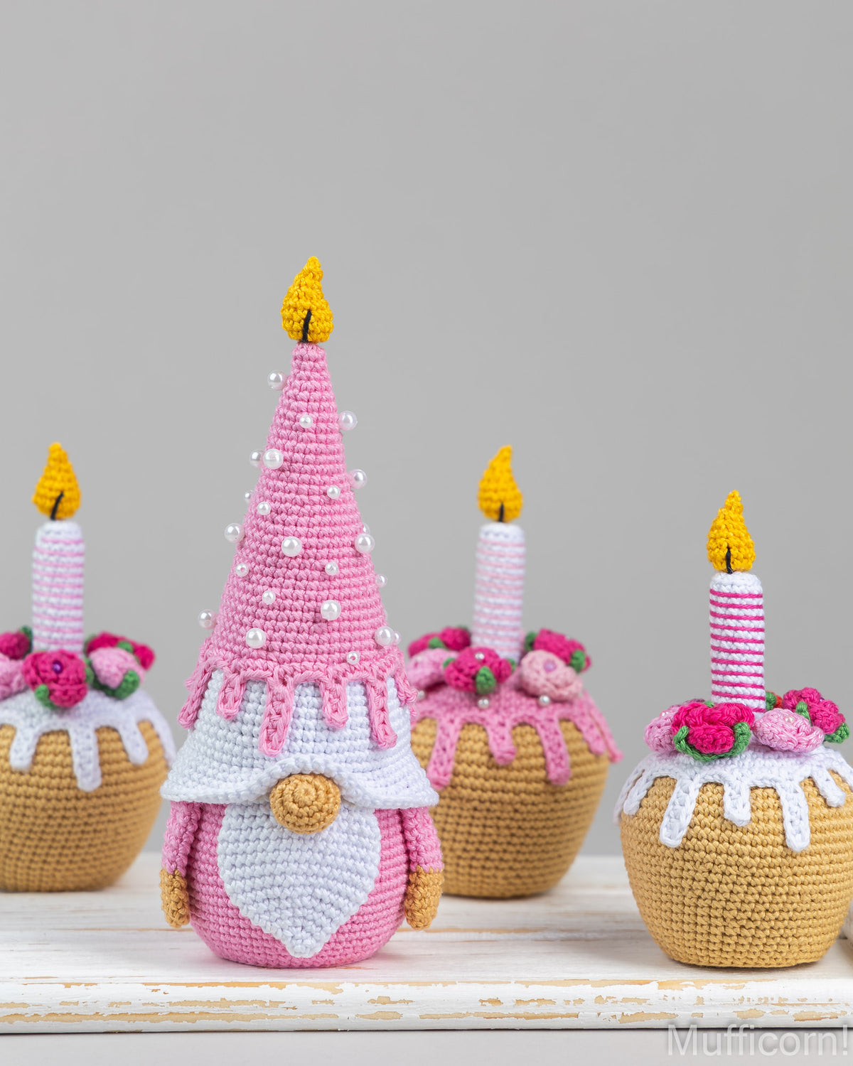 A crocheted gnome styled like a pink cupcake with a tall pointed hat, standing in front of three similar cupcake gnomes on a white wooden surface against a light gray background.