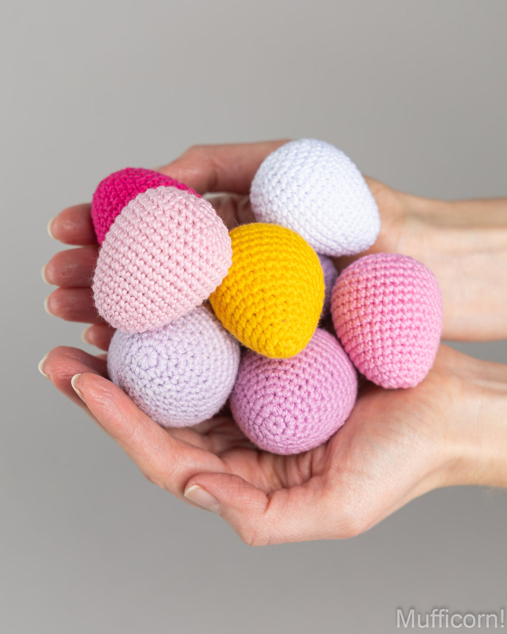 A pair of hands holding a cluster of crocheted yarn Easter eggs in pastel pink, lavender, white, yellow, and hot pink against a neutral gray background.
