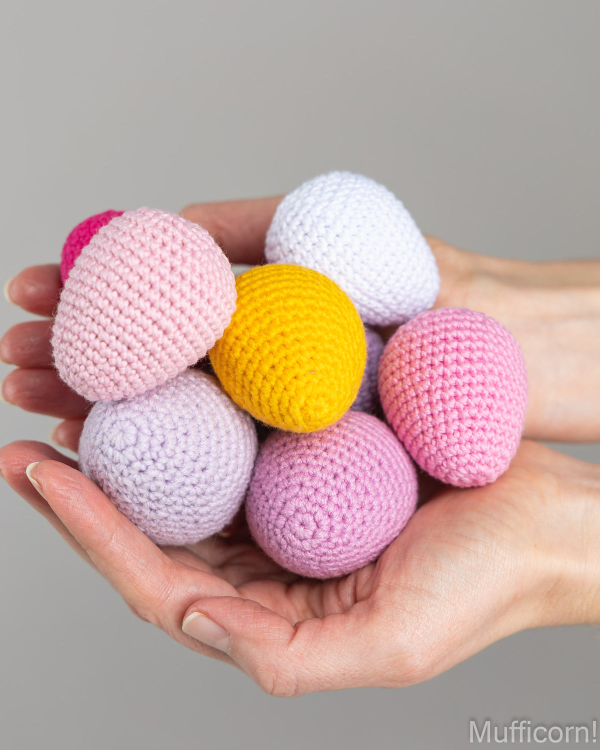 A pair of hands holding a cluster of crocheted Easter eggs in pastel colors, including pink, yellow, white, and lavender, against a solid gray background.
