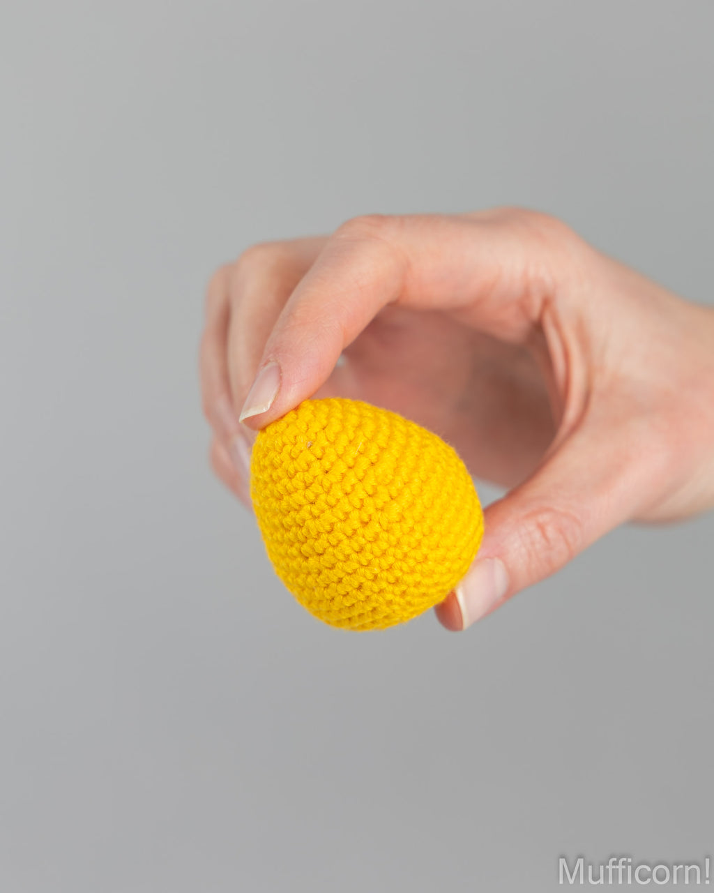 A hand holding a small, bright yellow crocheted egg-shaped ornament against a solid gray background.