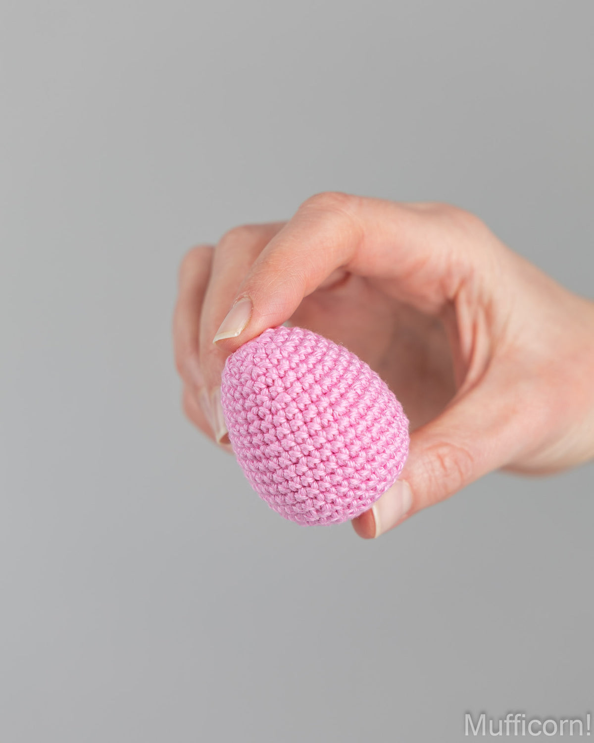 A hand holding a small pink crocheted egg-shaped ornament against a plain light gray background.