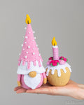 Two crocheted birthday gnome and muffin toys held in a person's hand against a plain gray studio background: a pink gnome with a tall cone-shaped hat decorated with white beads and a yellow flame, and a pink-and-white striped cupcake with a yellow flame, flowers, and white icing details.