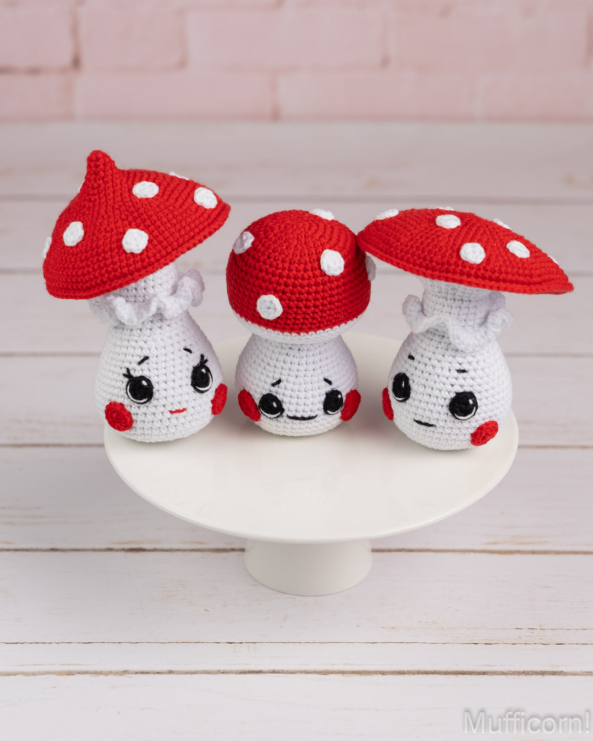 Three cute crocheted mushroom characters with red and white polka-dot caps and smiling faces, displayed on a white pedestal against a light pink brick wall and white wood floor.