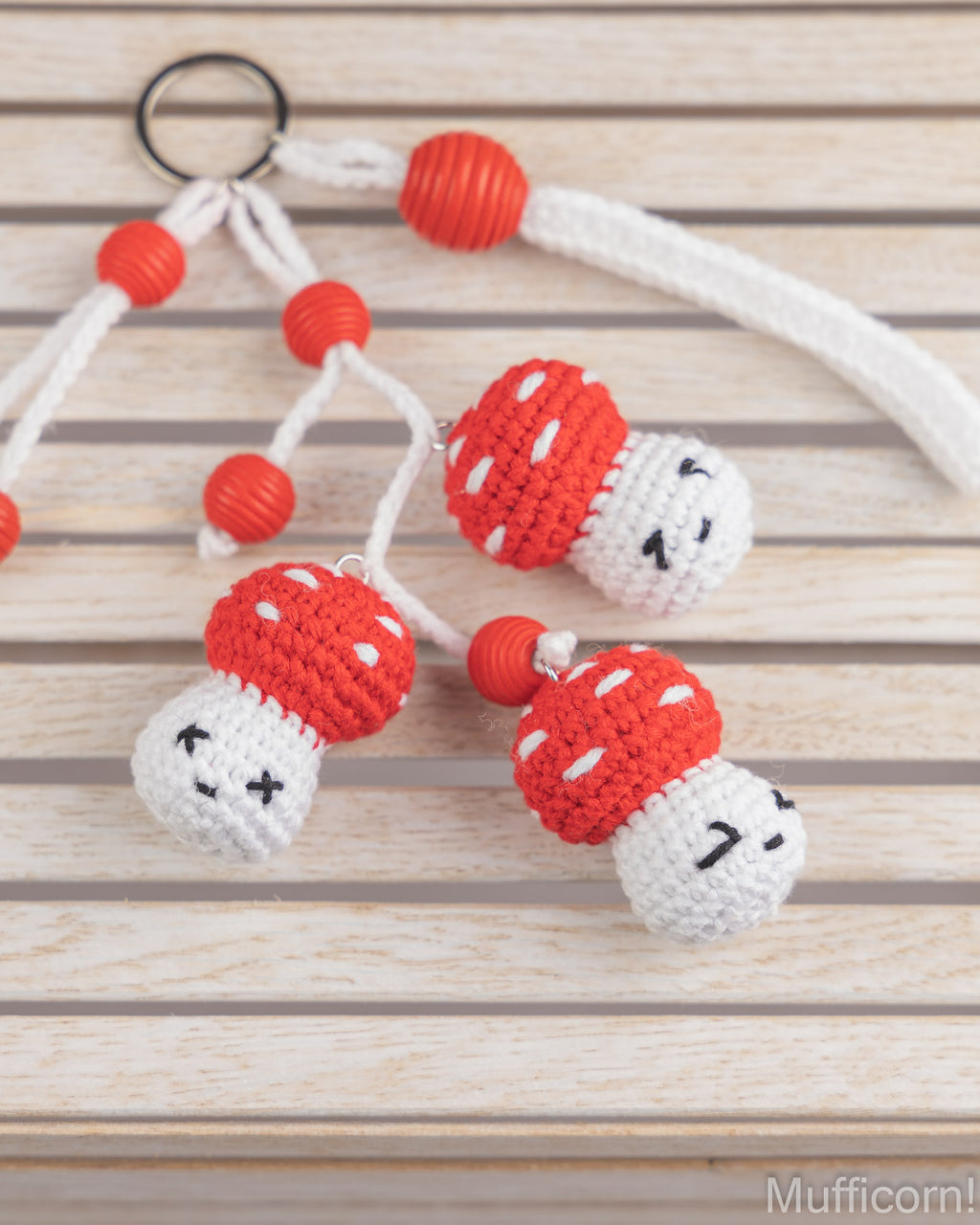 Three handmade crochet mushroom keychains with red and white bodies, white cords, and red beads, displayed on a light wood background.