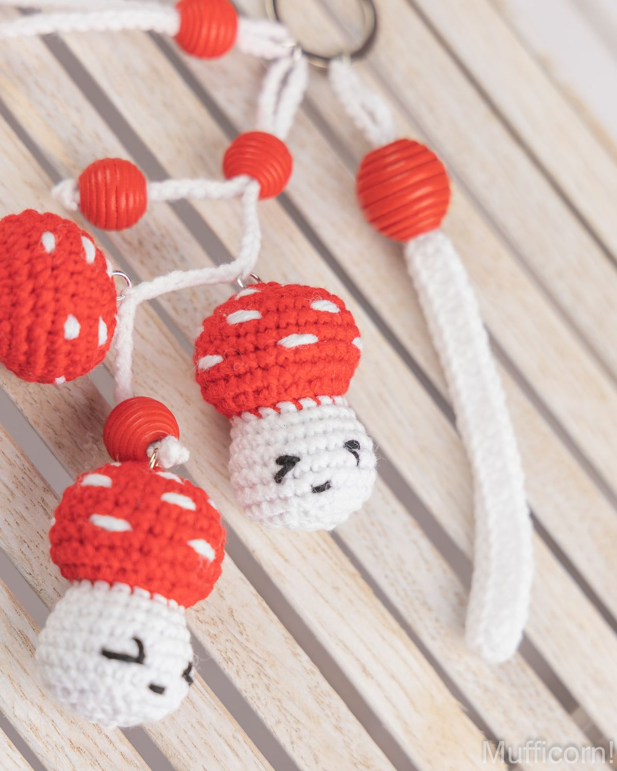 Three handmade crocheted mushroom keychains with red and white caps, white cords, and red beads, displayed on a light wood slatted surface.