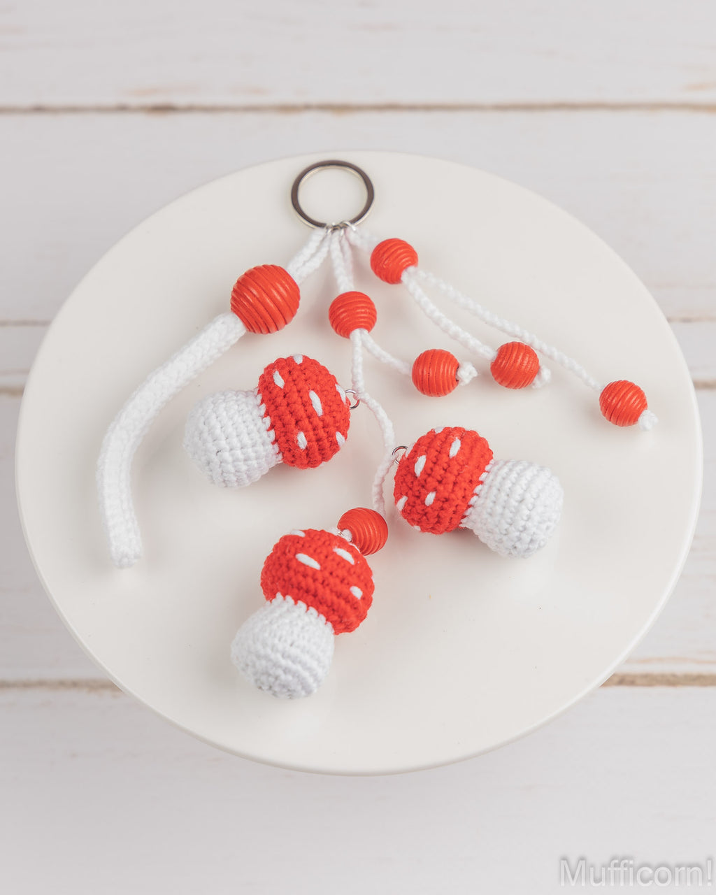 Three handmade crochet mushroom keychains with red and white bodies, white cords, and orange beads, displayed on a white plate over a light wood background.