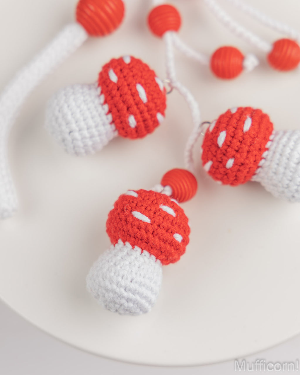 Crocheted mushroom-shaped jewelry charms with red caps, white stems, and small red beads, displayed on a white surface.