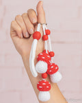 A hand holding three small crocheted mushroom keychains with red and white bodies, white loops, and red beads, against a soft pink brick wall background.
