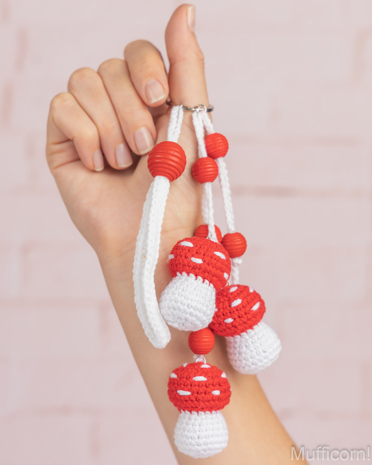 A hand holding three small crocheted mushroom keychains with red and white bodies, white loops, and red beads, against a soft pink brick wall background.