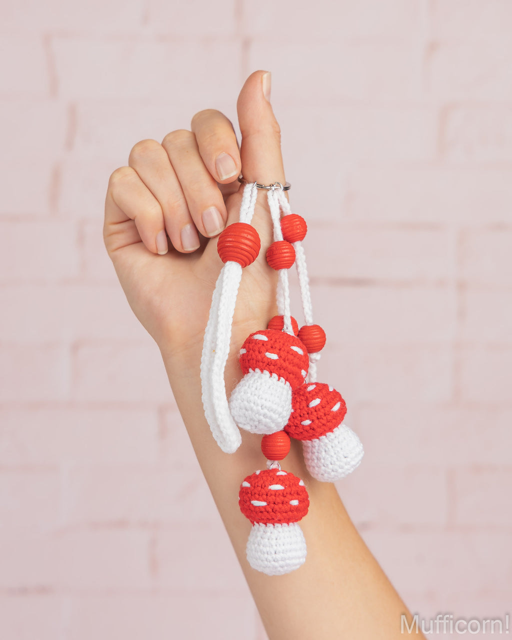 A hand holding a crocheted keychain featuring red and white mushroom charms with white cords and red beads, set against a light pink brick wall background.
