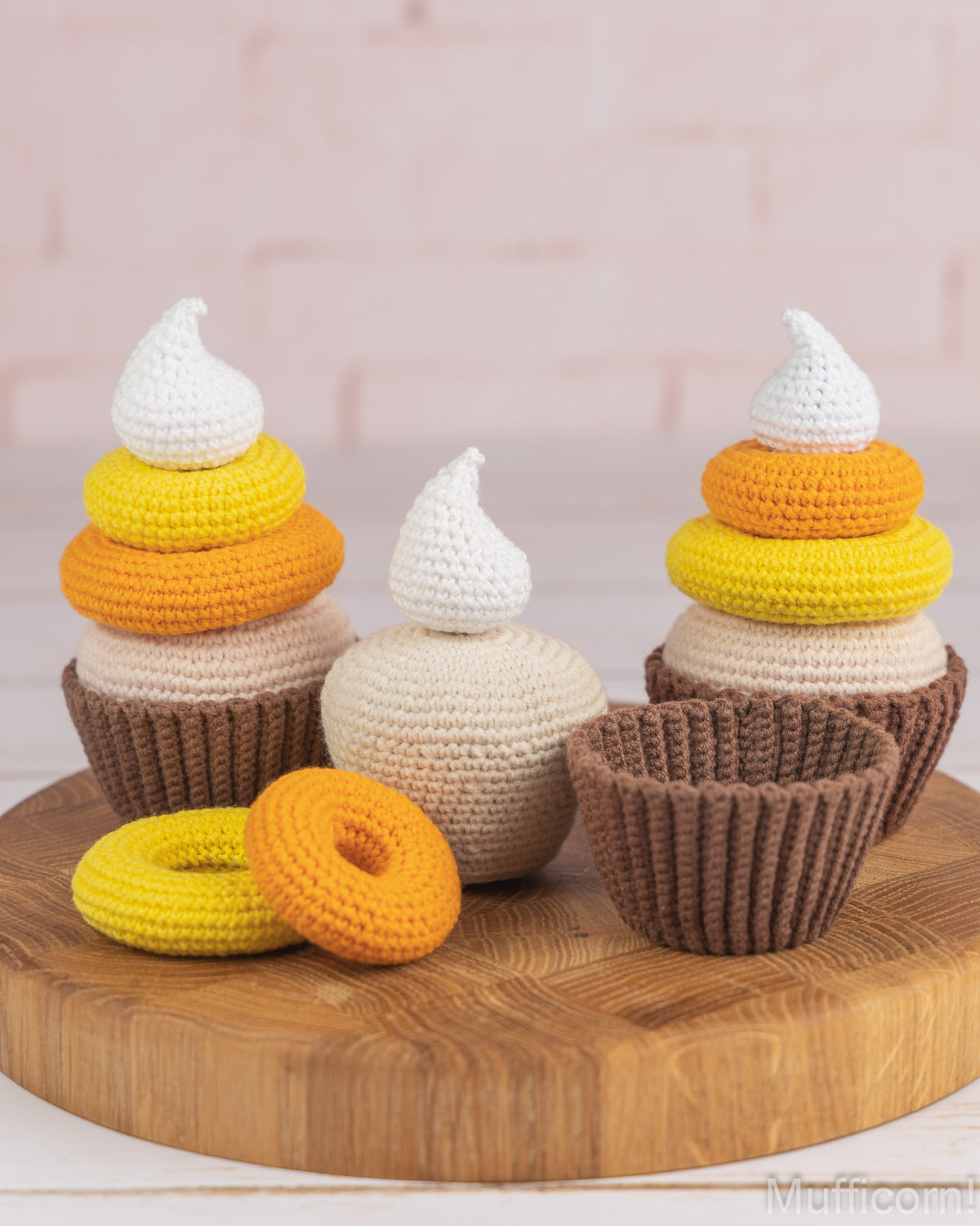 Three crocheted cupcake-shaped ornaments displayed on a round wooden board against a soft pink brick wall background. The set includes brown wrappers, yellow and orange rings, and white frosting tops.