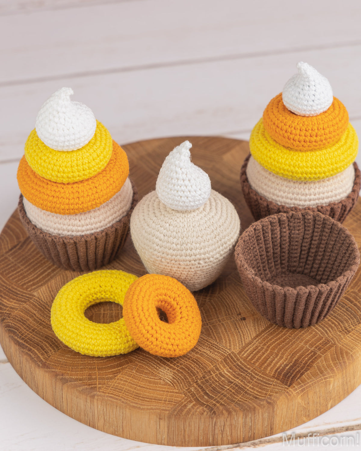 Crocheted cupcake and donut toys arranged on a round wooden tray against a light wood plank background. The set includes two cupcakes with white, yellow, and orange striped frosting-like tops, one cream-colored cupcake with a white top, one brown cupcake liner, and two yellow-orange donuts.