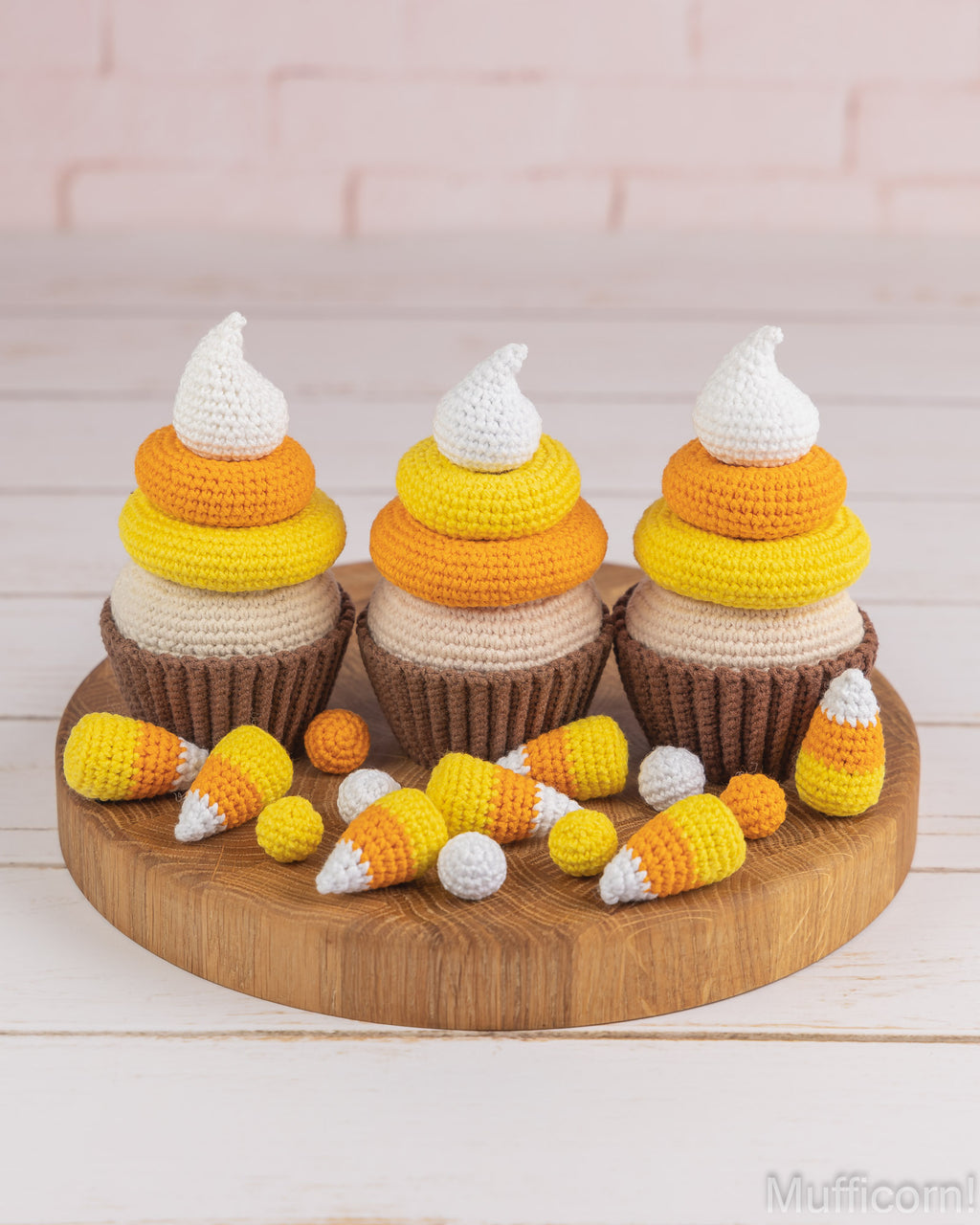 Three crocheted cupcakes styled like candy corn, arranged on a round wooden board with small crocheted candy corn pieces in front, against a pale pink brick wall and white wood surface.