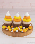 Three crocheted cupcakes styled like candy corn, arranged on a round wooden board with small crocheted candy corn pieces in front, against a pale pink brick wall and white wood surface.
