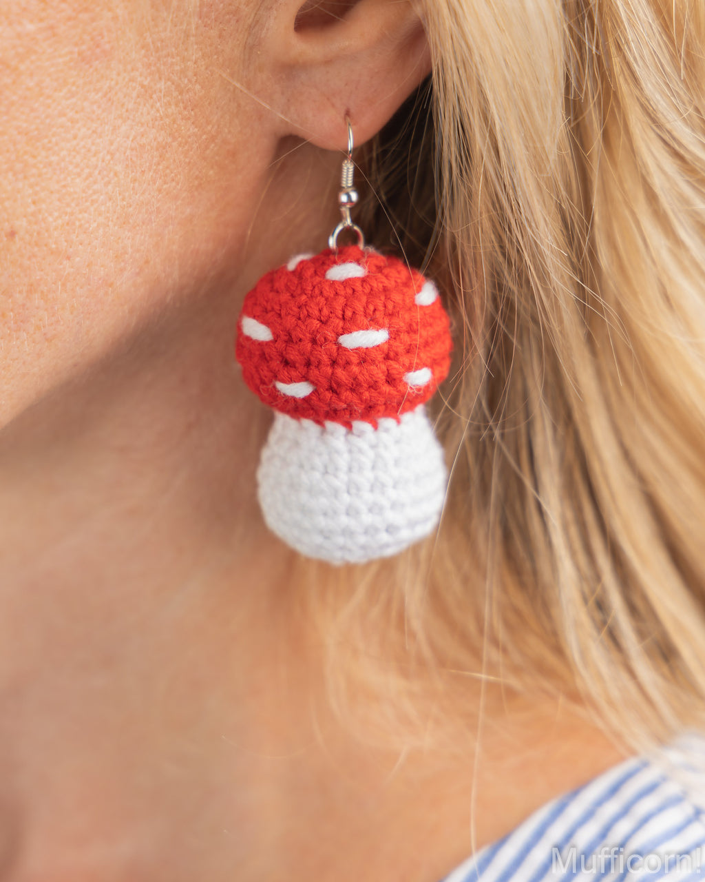 A close-up of a person's ear wearing a crocheted mushroom earring. The earring has a red top with white spots and a white bottom, hanging from a silver hook.