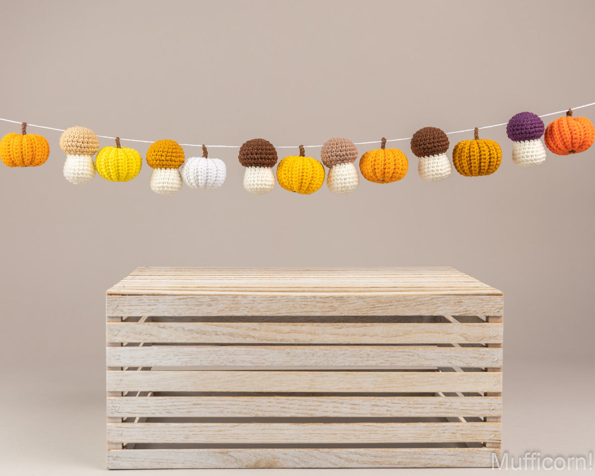 A string of crocheted pumpkin garland ornaments in orange, yellow, white, brown, and purple, displayed above a light wood slat crate on a neutral beige background.