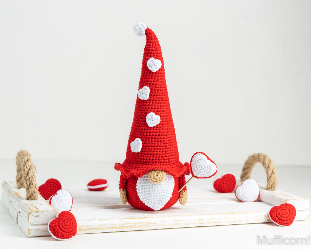 A crocheted gnome doll with a tall red hat decorated with white hearts, a white beard, and a small heart-shaped lollipop, displayed on a white wooden tray with red and white hearts around it.