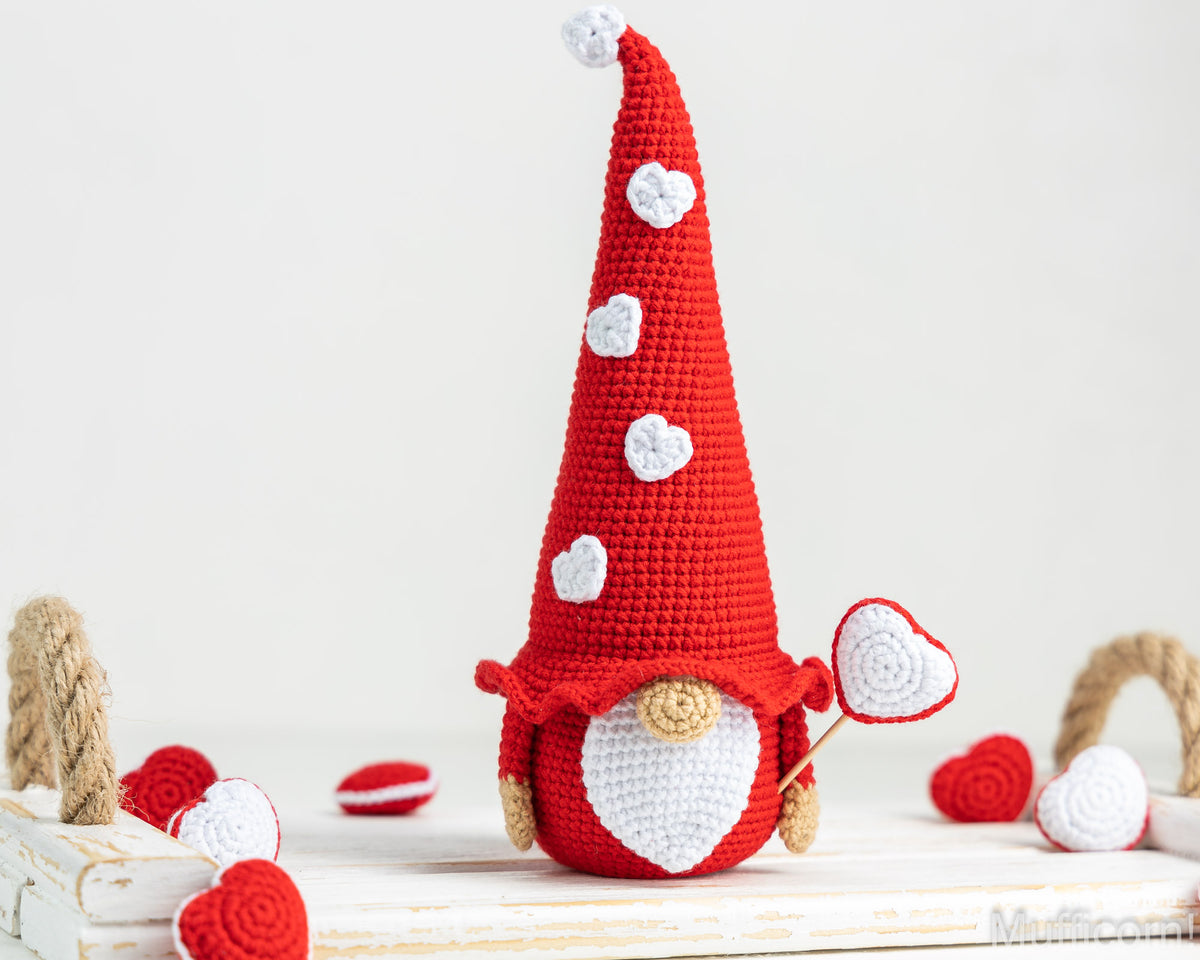 A crocheted gnome doll with a tall red hat decorated with white heart details, a white beard, and small tan hands, standing on a white wooden surface with red and white heart props around it.