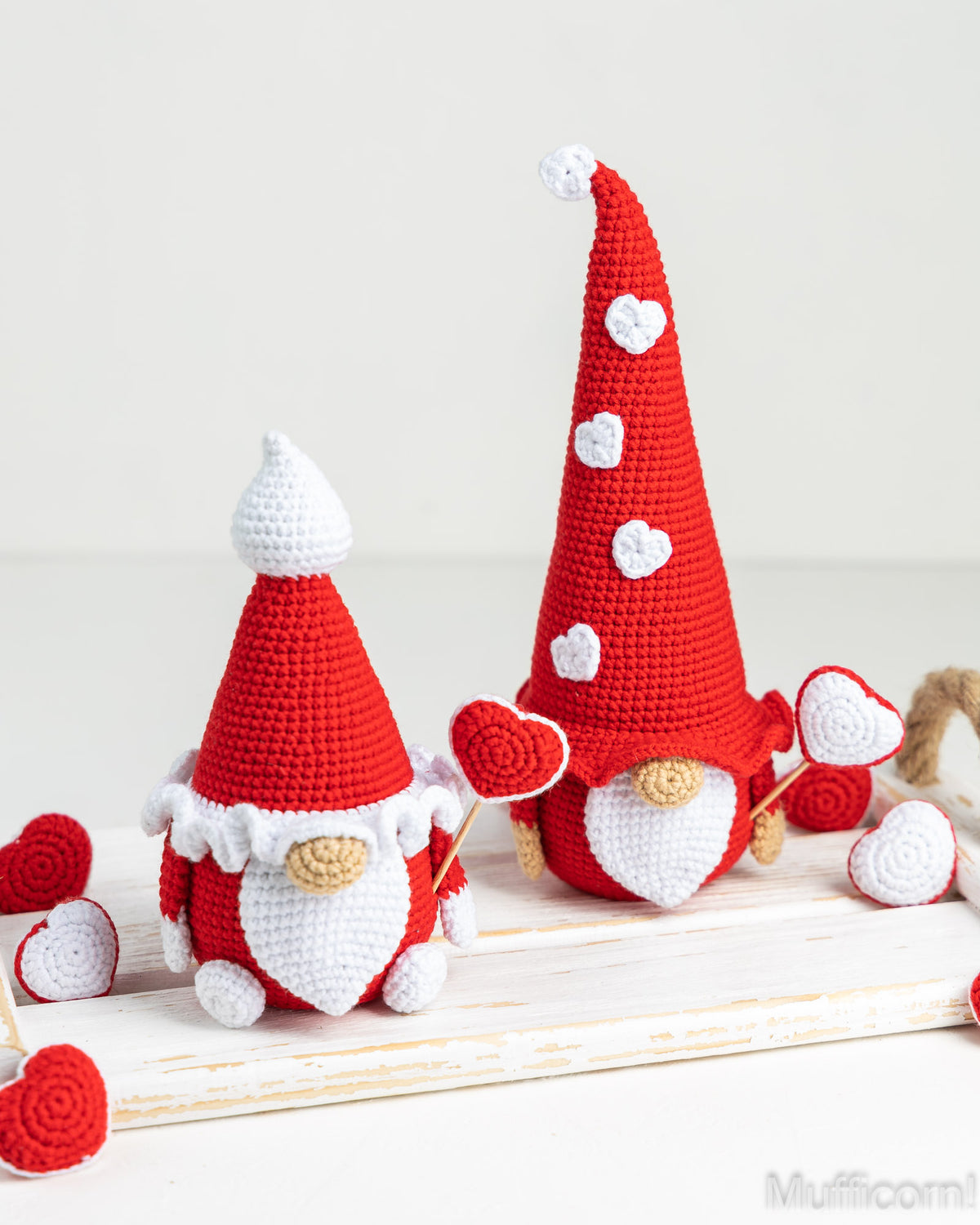 Two crocheted gnome dolls in red and white, styled with heart-shaped accessories on a white wooden surface against a light background.