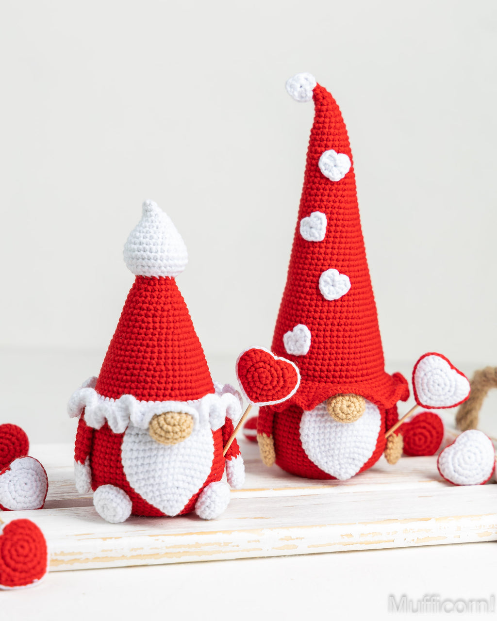 Two crocheted gnome figures in red and white with heart-shaped accessories, displayed on a light wooden surface against a soft off-white background.