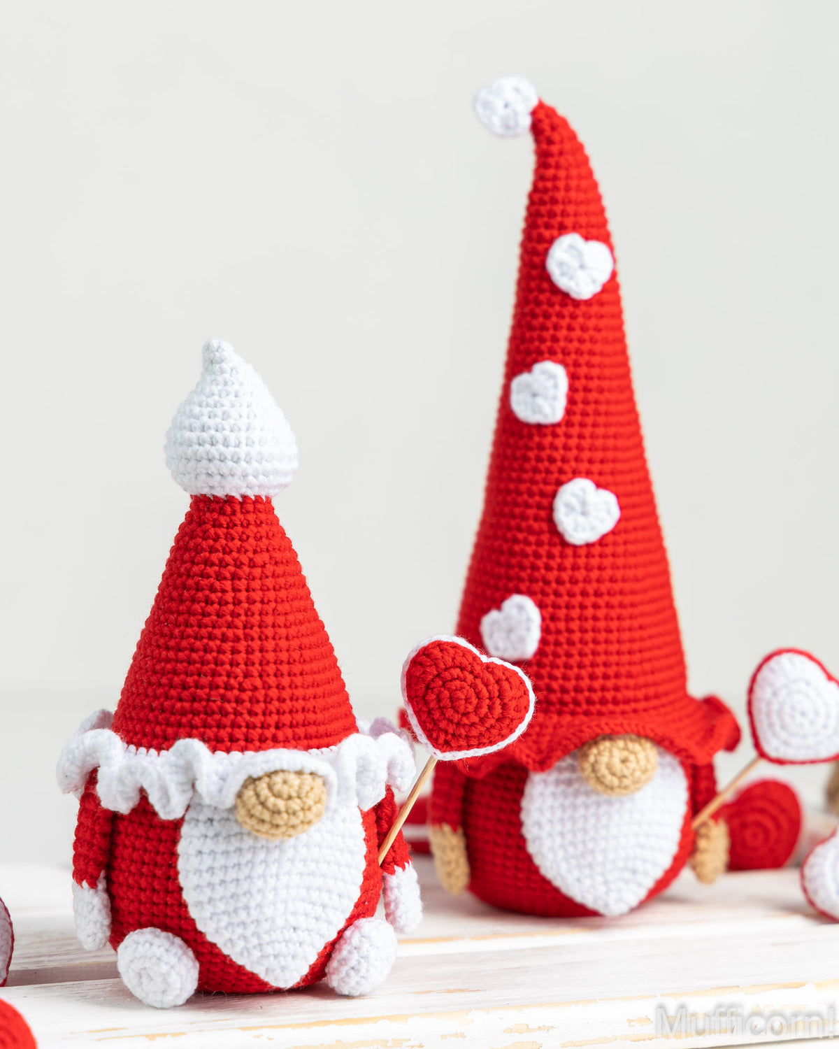 Two crocheted gnome decorations in red and white, styled on a light wooden surface against a soft off-white background. One small gnome holds a red-and-white heart-shaped lollipop, and a larger gnome stands behind it.