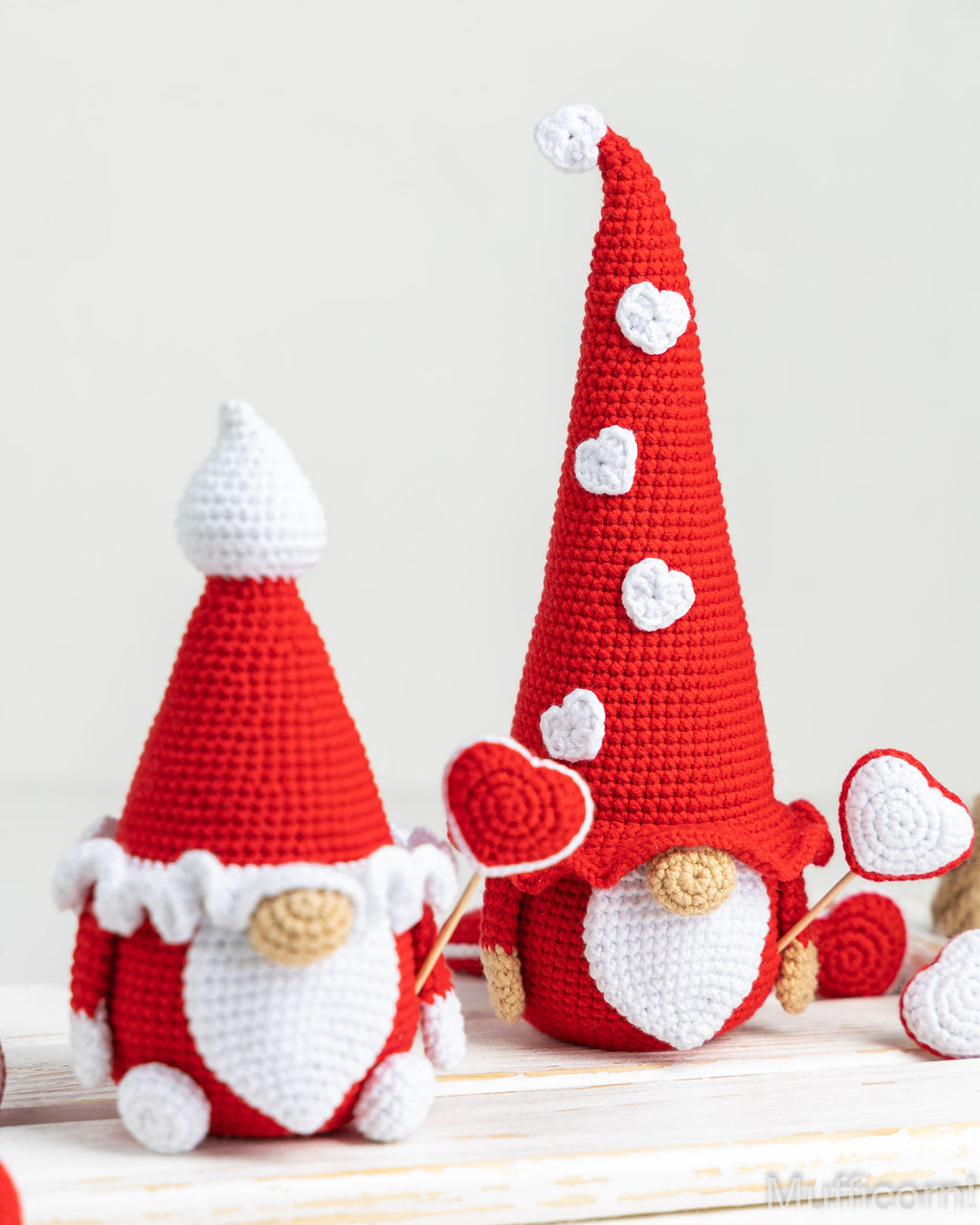 Two crocheted red and white gnomes with tall pointed hats, one holding a red heart-shaped lollipop and the other standing beside it, displayed on a light wooden surface against a soft off-white background.