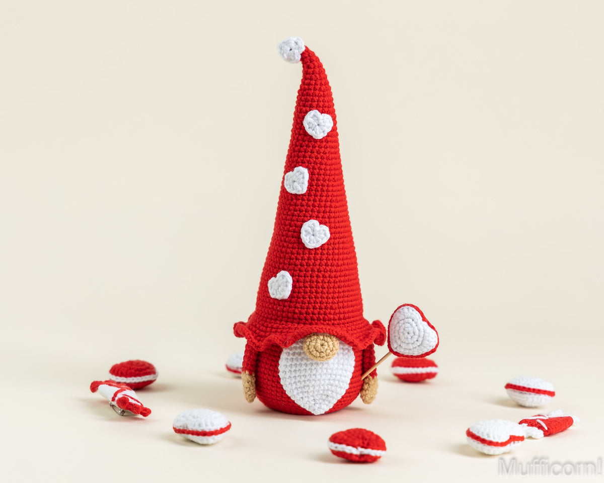 A crocheted red gnome doll with a tall pointed hat decorated with white hearts, standing on a light beige background with small red-and-white heart accessories scattered around it.