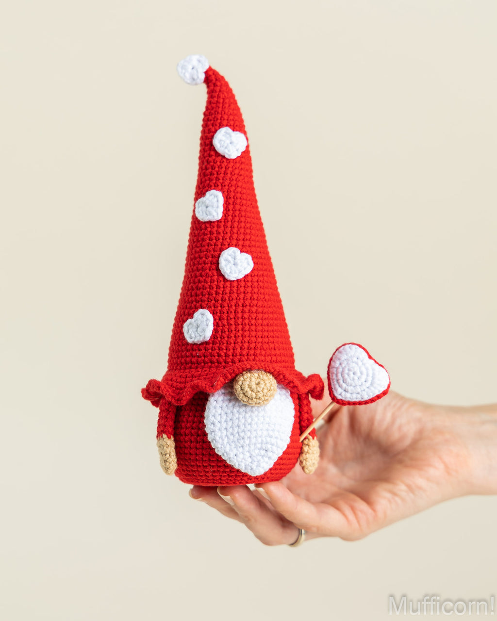 A crocheted gnome doll with a tall red hat decorated with white hearts, held in a person's hand against a plain beige background. The gnome has a white beard, tan hands and feet, and holds a small white heart.