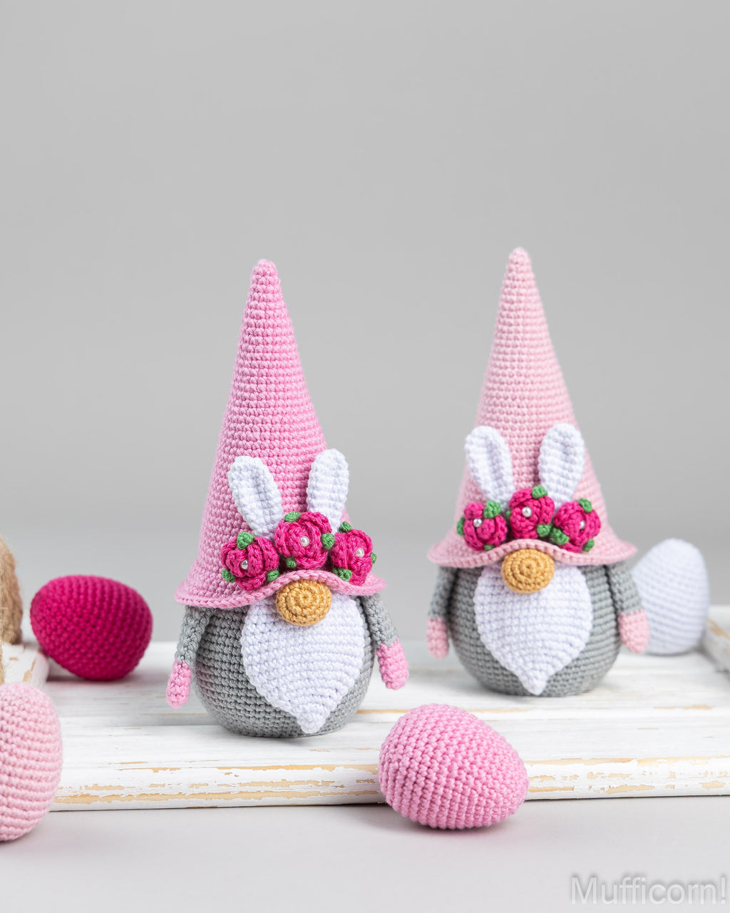 Two crocheted bunny gnomes with pink pointed hats, white bunny ears, pink flower crowns, and gray-and-white faces, displayed on a white wooden surface with pink and white yarn eggs around them.