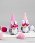 Two crocheted bunny gnomes with pink pointed hats, white bunny ears, pink flower crowns, and gray-and-white faces, displayed on a white wooden surface with pink and white yarn eggs around them.