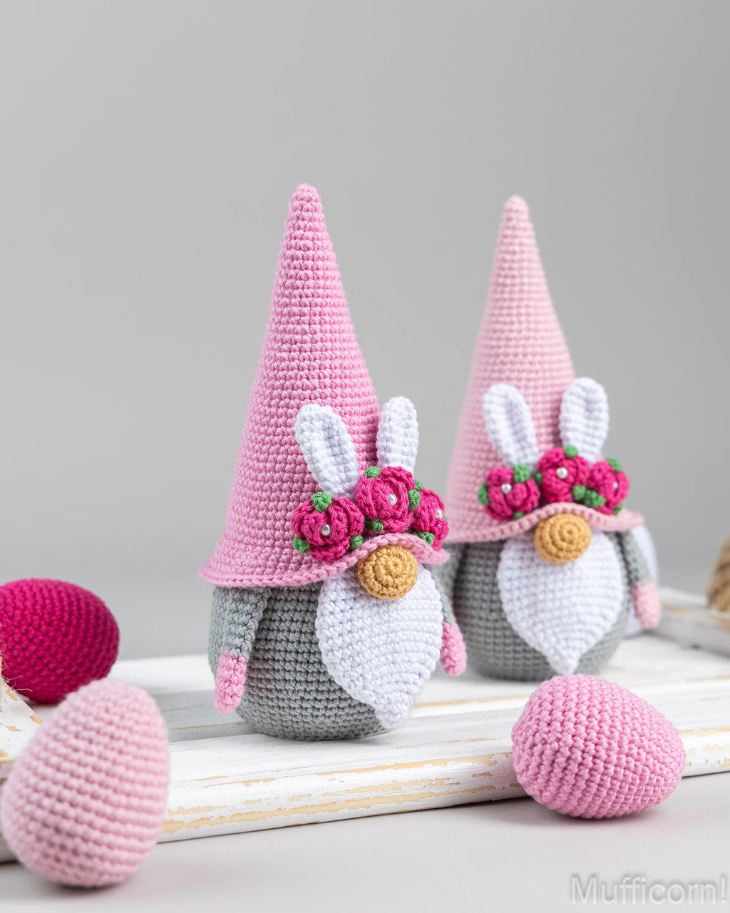 Two crocheted bunny gnomes with pink pointed hats, white beards, and pink flower crowns, displayed on a white wooden surface with pink and magenta egg-shaped props against a soft gray background.