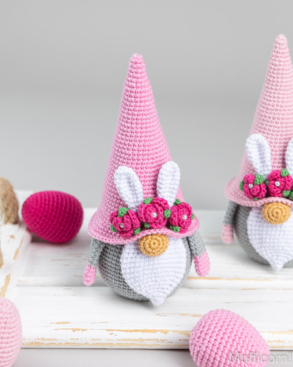 A crocheted Easter gnome doll with a tall pink hat, bunny ears, a white beard, and pink flower details, displayed on a white wooden surface with pink yarn eggs in the background.