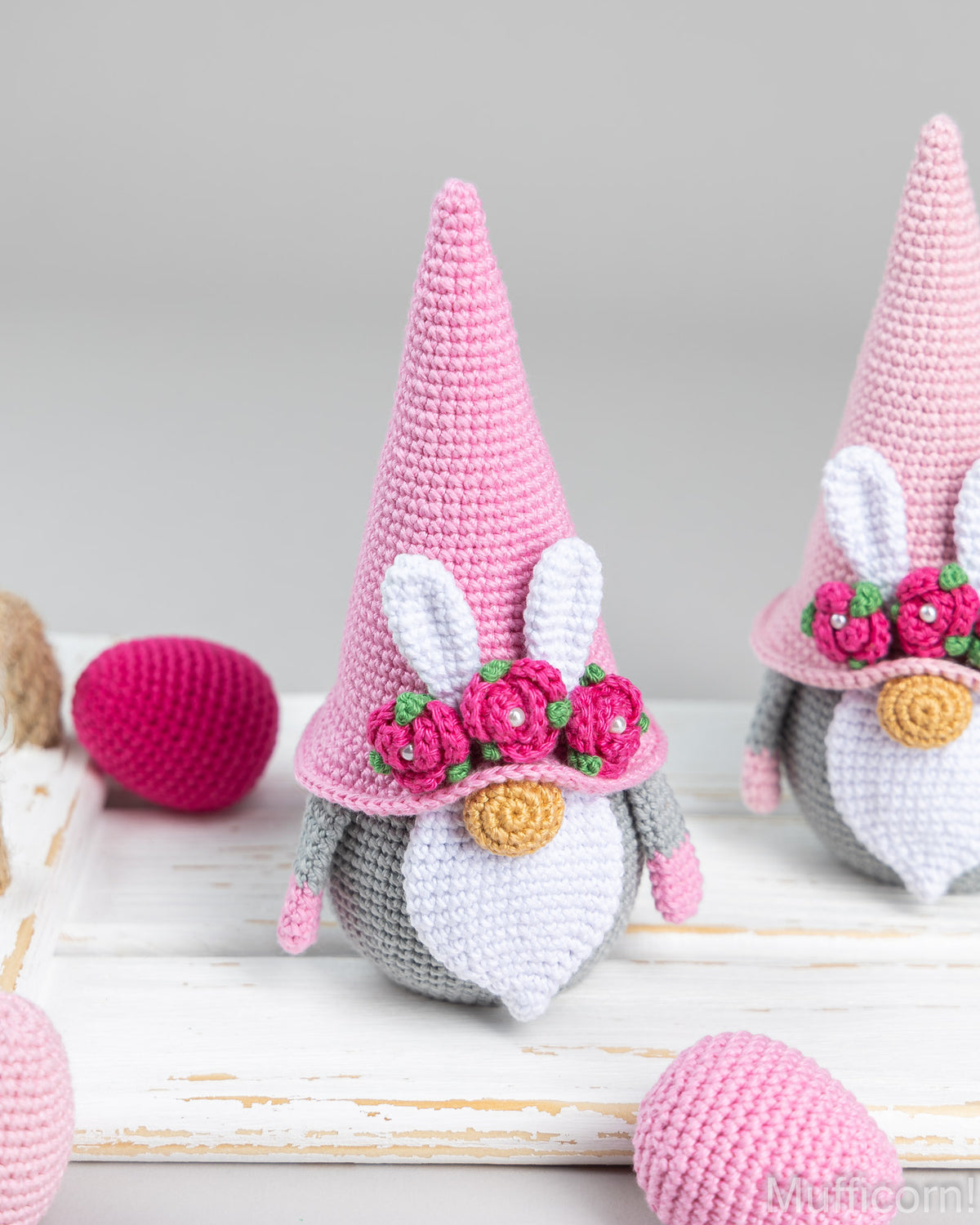A crocheted Easter gnome doll with a tall pink hat, bunny ears, a white beard, and pink flower details, displayed on a white wooden surface with pink yarn eggs in the background.