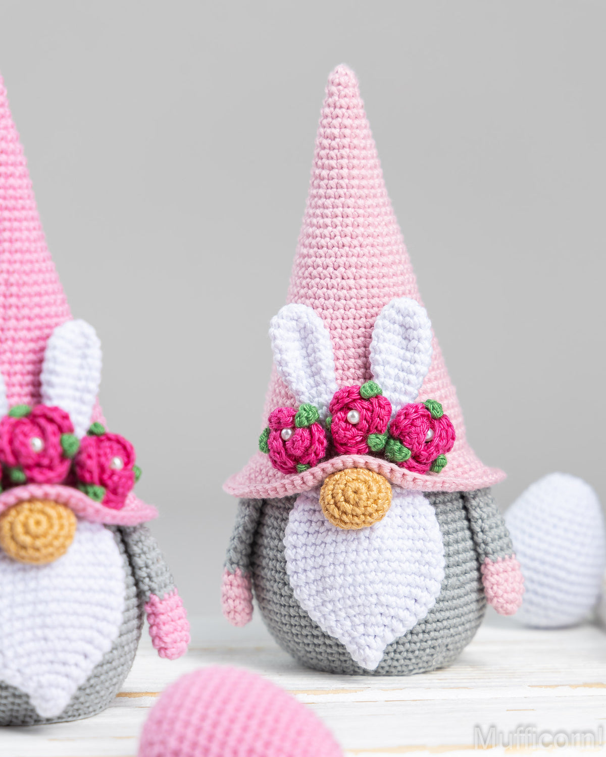 Crocheted bunny gnomes with pink and white hats, white beards, and pink flower crowns, shown on a light surface with soft gray background and pastel eggs.